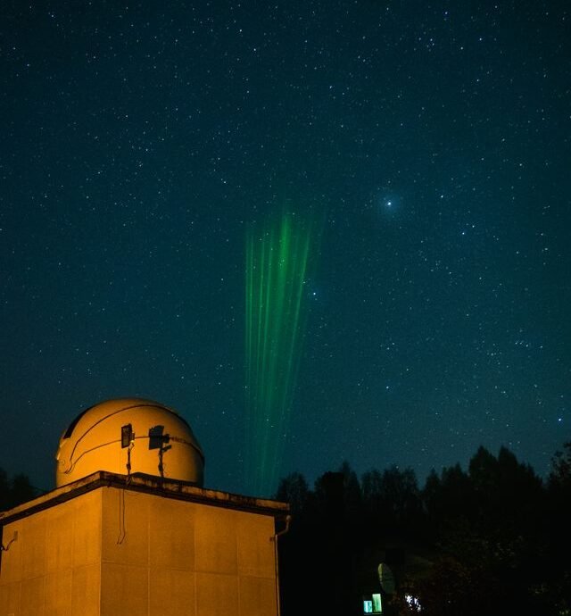 Observatory under starry sky with green laser beams pointing upwards.